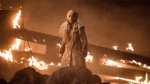 A personagem Daenerys Targaryen (Mãe dos Dragões) parece com um olhar desolador, beirando o choro em frente a uma grande destruição com muitos objetos pegando fogo e alguns corpos caídos bem a frente dela.